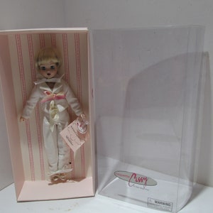 Puede incluir: Una muñeca blanca con cabello rubio que lleva un traje de marinero blanco con detalles rosas. La muñeca está en una caja de rayas rosas y blancas con las palabras "Coquette Crissy" en la caja.