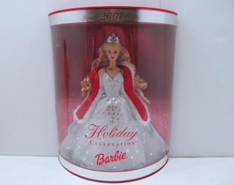 Może przedstawiać: Lalka Barbie Holiday Celebration w przezroczystej gablocie. Lalka ma na sobie srebrną suknię z gwiazdkami, czerwone pelerynę i tiarę. Na gablotce widnieje napis "Special 2001 Edition" i "Holiday Celebration Barbie."