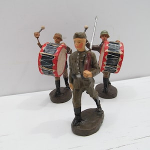 May include: Three toy soldiers, painted in military uniforms, holding drums and a sword. The soldiers are standing on a white surface.