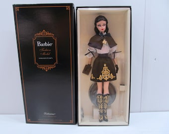 Puede incluir: Una muñeca Barbie de la colección Fashion Model Collection con un vestido marrón y dorado, una capa marrón y botas negras. La muñeca está de pie sobre una base negra y la caja tiene las palabras "Barbie Fashion Model Collection".