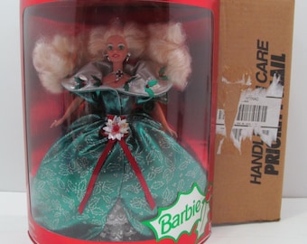 Może przedstawiać: Lalka Barbie w zielonej i srebrnej sukni z czerwoną wstążką i wzorem ostrokrzewu. Lalka znajduje się w przezroczystym plastikowym pudełku z czerwonym tłem. Pudełko zawiera napis "Wesołych Świąt" i "Edycja Specjalna".