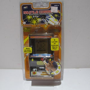 May include: A plastic package containing a handheld electronic game called "Missile Command: Classic Arcade". The game features a black and white LCD screen with a yellow border. The package is orange and yellow with a picture of the game on the front. The text "Real Arcade Sounds!" is printed on the package.