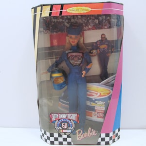 Puede incluir: Una muñeca Barbie vestida con un traje de carreras azul con un casco, sosteniendo una bandera de carreras. La muñeca está en una caja con un patrón de cuadros rojo, blanco y azul. La caja también presenta el texto "50th Anniversary NASCAR Barbie Collector's Edition".