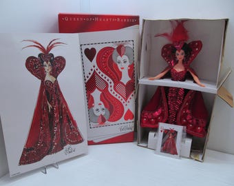 May include: Queen of Hearts Barbie doll in a red and white dress with a heart-shaped bodice and a red cape. The doll is holding a playing card with the Queen of Hearts on it. The doll is packaged in a box with a red and white design.