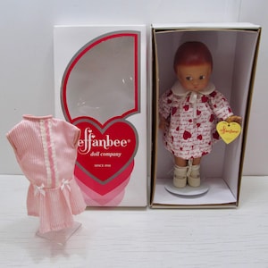 Puede incluir: Muñeca Effanbee en una caja, con un vestido con estampado de corazones y zapatos blancos. Un vestido a rayas rosas y blancas se muestra junto a la caja. La caja presenta el logotipo de Effanbee y un diseño de corazón.