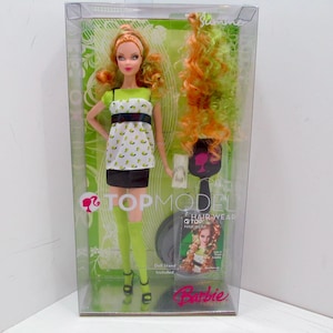 Puede incluir: Una muñeca Barbie Top Model con ropa estampada en verde y blanco, cabello naranja y una peluca verde y negra. La muñeca está empaquetada en una caja de plástico transparente con las palabras "Top Model" y "Barbie" en ella.
