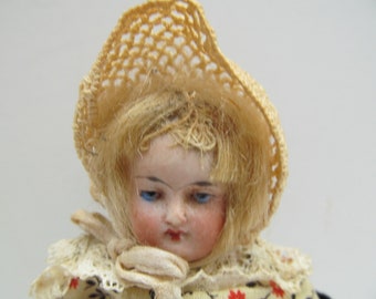 May include: A porcelain doll with blonde hair wearing a white crocheted hat and a white lace collar. The doll has blue eyes and rosy cheeks.