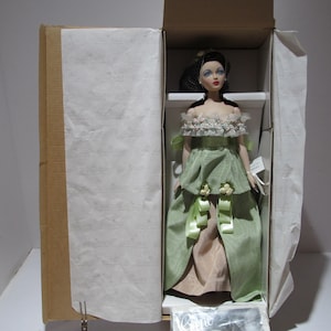 Könnte beinhalten: Eine Porzellanpuppe in einem grün-beigen Kleid mit Spitzendetails. Die Puppe befindet sich in einer Schachtel mit weißem Seidenpapier.