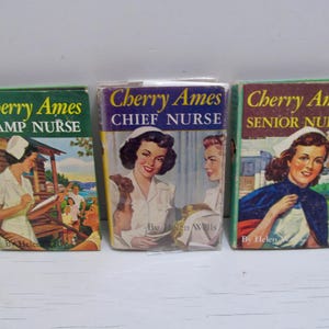May include: Three vintage Cherry Ames books. The titles are "Camp Nurse", "Chief Nurse", and "Senior Nurse". The covers feature illustrations of nurses in various settings. The books are in good condition, with visible wear and tear.