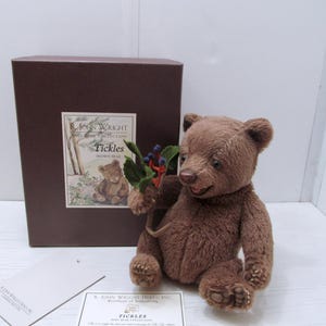 May include: A brown teddy bear named "Tickles" sits next to a brown box with a label. The bear holds a small sprig of berries and leaves. The box has a label with the bear's name and a picture of a bear.
