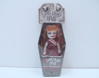 Puede incluir: Una muñeca Living Dead Dolls en un paquete con forma de ataúd. La muñeca tiene el pelo rojo, ojos azules y una boca ensangrentada. Viste un vestido rojo y una banda que dice "Danse Macabre". El paquete tiene las palabras "Living Dead Dolls".