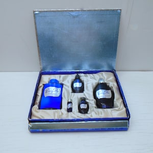 May include: A vintage perfume set in a blue box. The set contains several blue glass bottles of different sizes, each with a white label. The box is lined with beige fabric and has a silver lid.