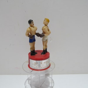 May include: A vintage toy boxing game with two plastic figures, one with black hair and one with blonde hair. The figures are wearing boxing gloves and shorts. The base of the game is red and has the words "Hit 'n Miss" printed on it.