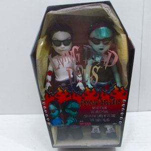May include: Two Living Dead Dolls in a coffin-shaped box. The dolls are dressed in black and white clothing and have black sunglasses. The box has the text "Living Dead Dolls" and "Psycho-Billies" on it.