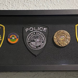 May include: A framed display of three embroidered police patches from Hialeah Gardens, Florida. The patches are in a black frame with a black background. The patches are all gold and blue with the words "Police" and "Hialeah Gardens, Florida" on them. The patches are all different sizes and shapes.