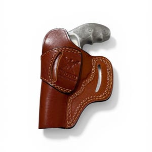 Puede incluir: Una funda de cuero marr&oacute;n con una pistola plateada. La funda est&aacute; cosida con hilo marr&oacute;n claro y presenta un logotipo estampado. La empu&ntilde;adura de la pistola es visible, contrastando con el color de la funda.