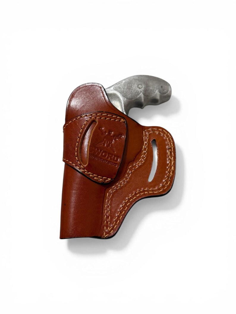 Puede incluir: Una funda de cuero marr&oacute;n con una pistola plateada. La funda est&aacute; cosida con hilo marr&oacute;n claro y presenta un logotipo estampado. La empu&ntilde;adura de la pistola es visible, contrastando con el color de la funda.