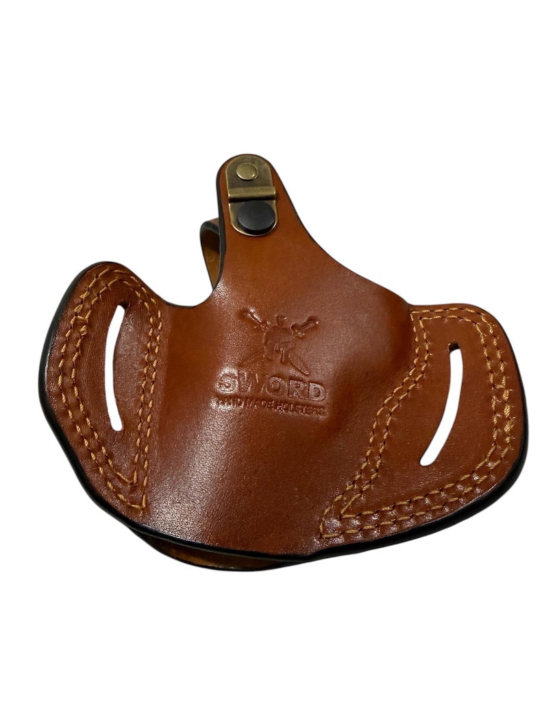 May include: A brown leather holster with visible stitching and the text "SWORD HAND MADE HOLSTERS". The holster has two oval cutouts and a metal clip. The leather is a medium brown color.