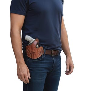 May include: A brown leather holster with a gun inside, worn on a brown belt over blue jeans. The holster is stitched and appears to be custom-made. The person is wearing a navy blue t-shirt.