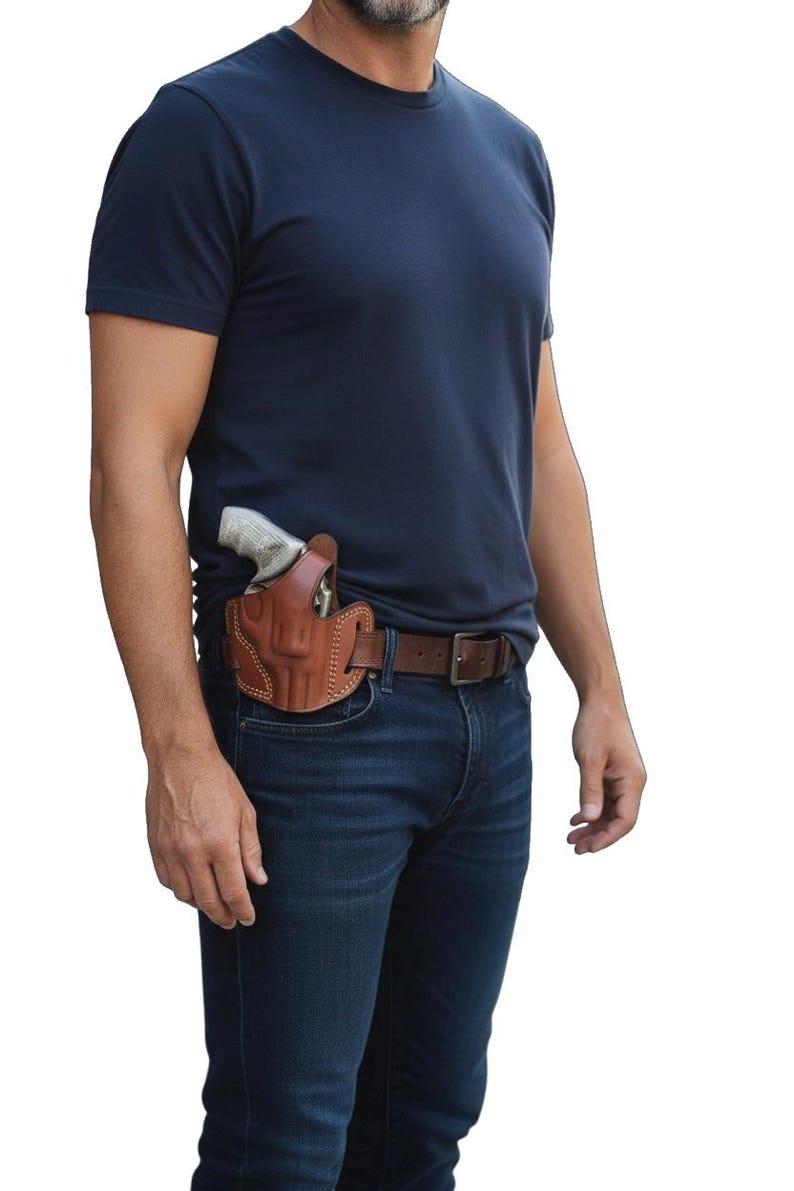 May include: A brown leather holster with a gun inside, worn on a brown belt over blue jeans. The holster is stitched and appears to be custom-made. The person is wearing a navy blue t-shirt.