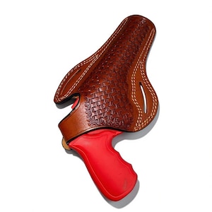 Puede incluir: Una funda de cuero marrón con un patrón tejido y costuras blancas. La funda contiene una pistola roja. El diseño presenta una forma curva con aberturas para el cinturón y un ajuste seguro para el arma de fuego.