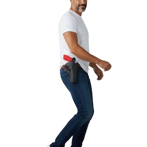 Pode incluir: Uma coldre de couro preto com uma arma de fogo vermelha está preso à cintura de uma pessoa vestindo uma camiseta branca e jeans azuis. O coldre foi projetado para ser usado na cintura. A pessoa está usando tênis brancos.