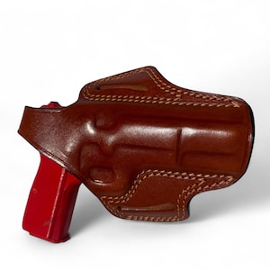 May include: A brown leather holster with detailed stitching, holding a red handgun. The holster is designed to fit the gun's shape, with visible seams and a secure fit. The image is set against a white background.
