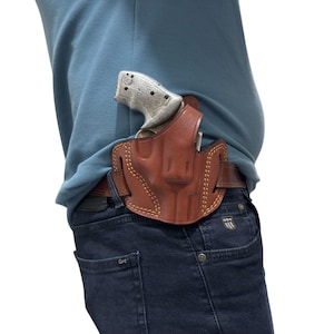 May include: A brown leather holster with a stitched design, holding a silver and black handgun. The holster is worn on a brown belt, secured to dark blue jeans. The image is set against a white background.