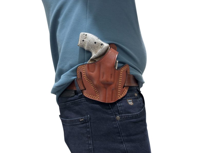 May include: A brown leather holster with a stitched design, holding a silver and black handgun. The holster is worn on a brown belt, secured to dark blue jeans. The image is set against a white background.
