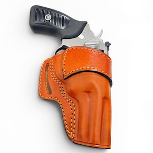 May include: A silver revolver with a black textured grip is partially inserted into a tan leather holster. The holster features prominent orange stitching along its edges and a moulded shape that contours to the frame of the firearm.