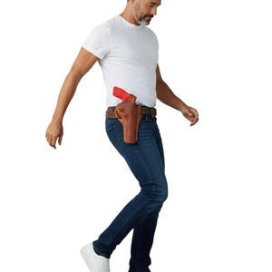 Op de afbeelding: Een bruin leren holster met een rood pistool is bevestigd aan een bruine riem. Het holster heeft een geweven patroon. De persoon draagt een wit t-shirt, een blauwe spijkerbroek en witte sneakers.