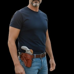 Puede incluir: Un hombre con una camiseta azul marino y vaqueros azules. Una funda de cuero marr&oacute;n con un rev&oacute;lver plateado est&aacute; sujeta al cintur&oacute;n de cuero marr&oacute;n. El fondo es negro.
