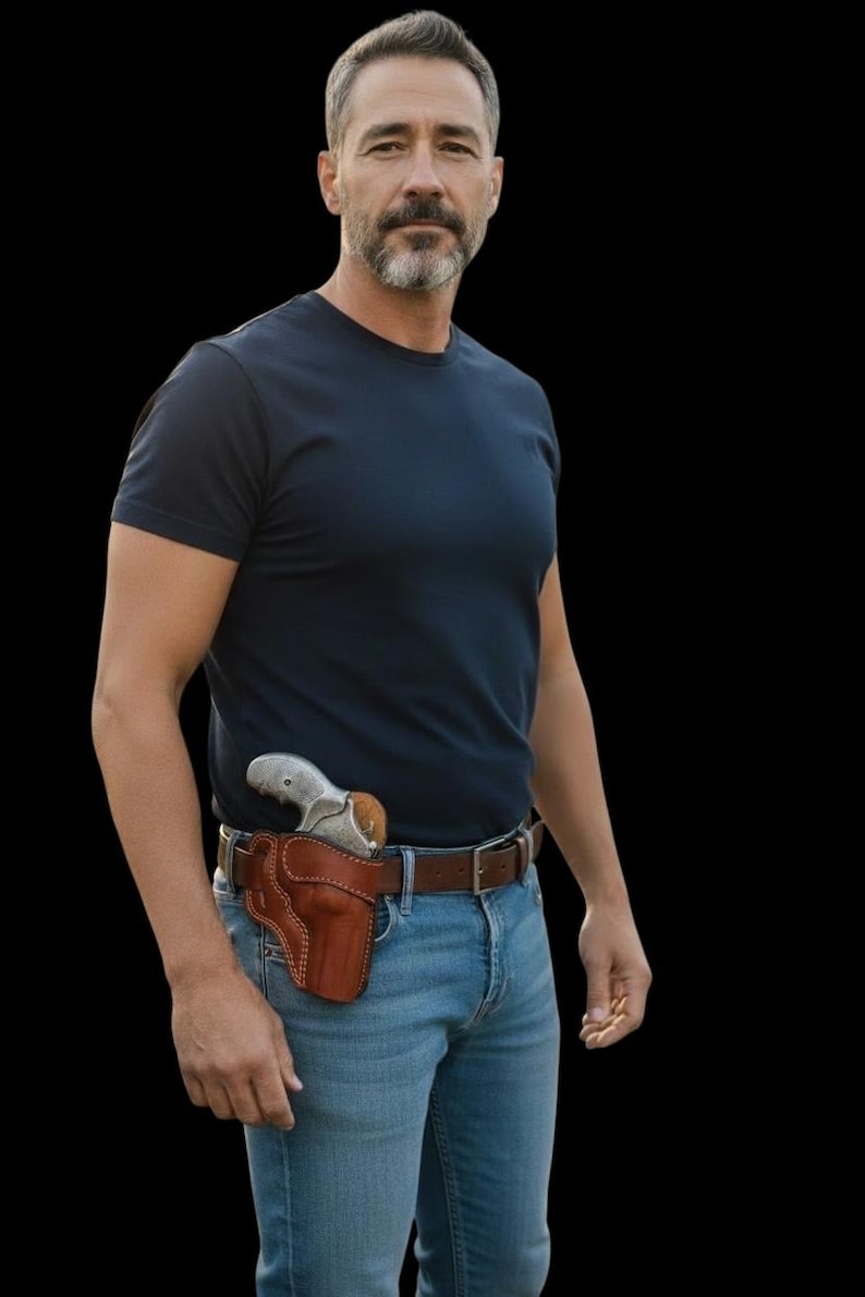 Puede incluir: Un hombre con una camiseta azul marino y vaqueros azules. Una funda de cuero marr&oacute;n con un rev&oacute;lver plateado est&aacute; sujeta al cintur&oacute;n de cuero marr&oacute;n. El fondo es negro.