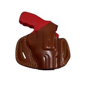 May include: A brown leather holster with a red handgun grip. The holster is stitched with tan thread and has a curved shape. The handgun grip is visible at the top of the holster.