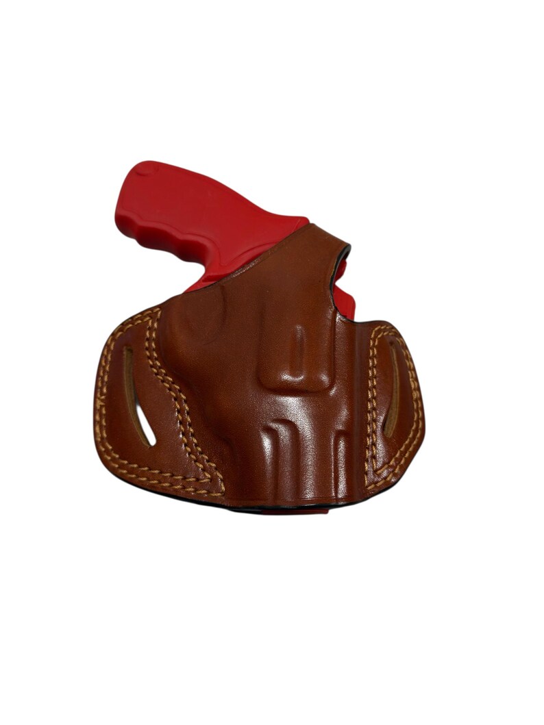 May include: A brown leather holster with a red handgun grip. The holster is stitched with tan thread and has a curved shape. The handgun grip is visible at the top of the holster.
