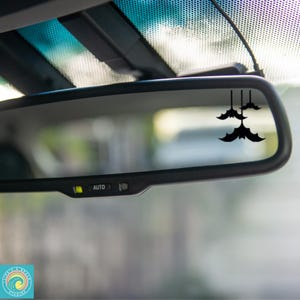 May include: Black bat car decal hanging from a car's rearview mirror. The decal features three bats of varying sizes, perfect for Halloween or a spooky aesthetic. The mirror has an "AUTO" button.