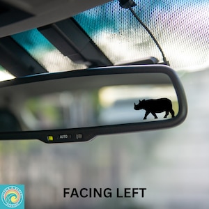 May include: A black silhouette of a rhinoceros facing left is visible in the rearview mirror of a car. The mirror is labeled "AUTO". The text "FACING LEFT" is visible below the mirror.
