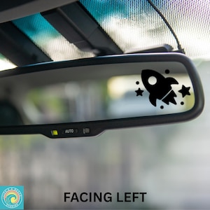 May include: A black rocket ship and star decal is affixed to a car's rearview mirror. The decal is on the right side of the mirror, with the text "FACING LEFT" below the mirror. The decal is a fun, decorative item for a vehicle.