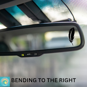 May include: A black feather sticker on a rearview mirror in a car. The sticker is positioned to the right of the mirror. The text "BENDING TO THE RIGHT" is visible below the mirror.