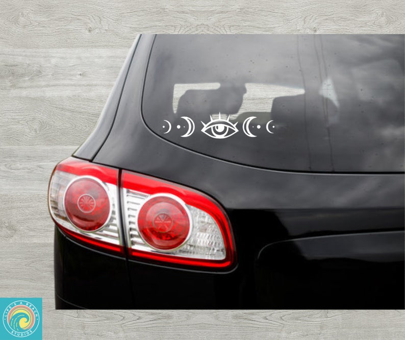 May include: A black car with a white sticker on the rear window. The sticker depicts a stylised eye with crescent moons on each side.