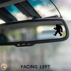May include: A black Bigfoot silhouette sticker is affixed to a car's rearview mirror. The Bigfoot figure is walking and carrying a golf club over its shoulder. The text "FACING LEFT" is at the bottom of the image.