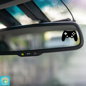May include: A black controller sticker is affixed to a car's rearview mirror. The sticker is a silhouette of a video game controller, featuring buttons and directional controls. The mirror also has an "AUTO" button.