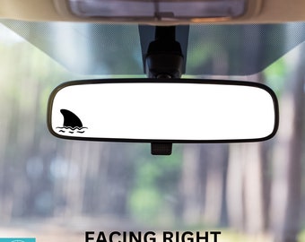 May include: A black shark fin sticker on a rearview mirror in a car. The sticker is facing right and says "FACING RIGHT" below it.