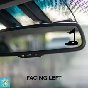 May include: A black car rearview mirror with a black golf flag sticker on the glass. The sticker shows a golf flag with a golf ball on a green.