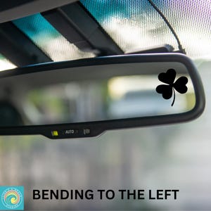 May include: A black car rearview mirror with a black shamrock sticker on the glass. The mirror has an "AUTO" button on the bottom. The text "BENDING TO THE LEFT" is visible below the mirror.