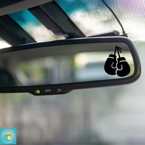 May include: A black silhouette of a pair of boxing gloves hanging from a string, visible in the reflection of a rearview mirror in a car.