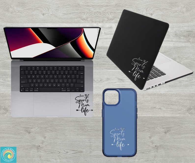May include: A blue phone case with a white design that says "Live that Sports Mom Life" with an arrow pointing to the right. The case is on a wooden surface next to a laptop with a similar design on the back.