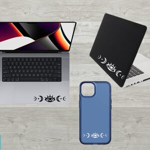 May include: A laptop, phone case, and laptop skin with a white eye and crescent moon design. The laptop is silver and has a black keyboard. The phone case is blue and the laptop skin is black.