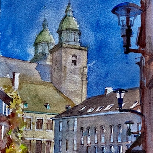 May include: Watercolor painting of a European cityscape with a bright blue sky. The artwork depicts buildings with arched windows, a church with two green-domed towers, and street lamps. The style is loose and impressionistic.