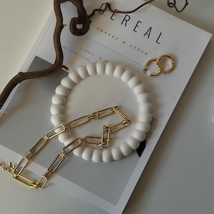 May include: A gold chain necklace with rectangular links on a white ceramic dish with a textured surface. The dish is on top of a magazine with the text "Cereal Travel & Style".